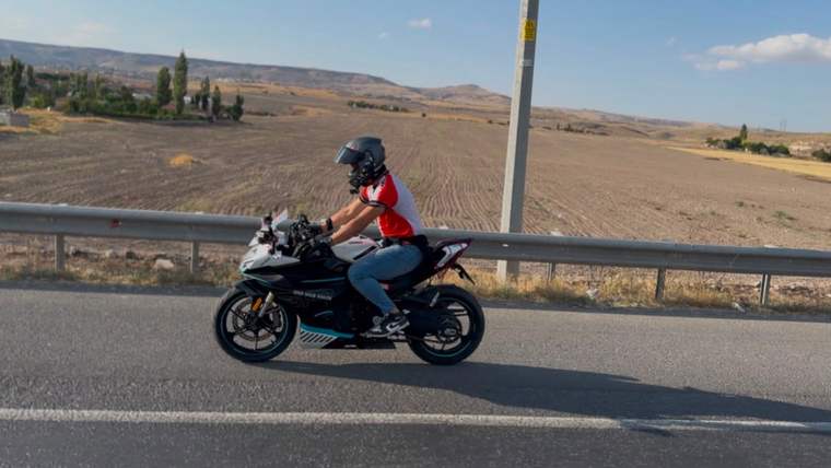 Kasklı bir kişi, kırmızı tişörtle motosiklet üzerinde kırsal yolda ilerlerken görülüyor.