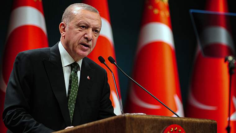 Kürsüde konuşan bir kişi, arkasında Türk bayraklarıyla birlikte mikrofonlara hitap ediyor.