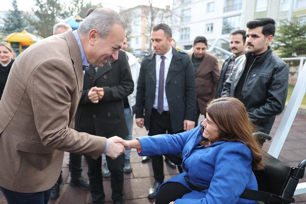 Açık alanda tekerlekli sandalyede oturan bir kadın, çevresindeki insanların önünde bir erkekle tokalaşıyor.