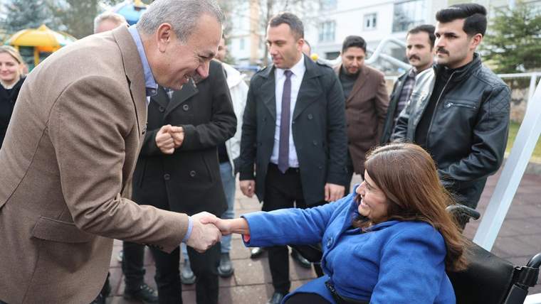 Açık alanda tekerlekli sandalyede oturan bir kadın, çevresindeki insanların önünde bir erkekle tokalaşıyor.