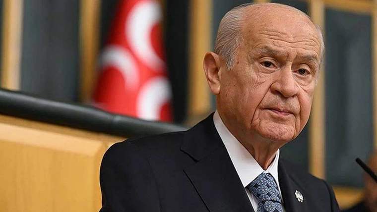 Kürsüde konuşan bir kişi, arka planda Türk bayrağı ve ahşap paneller önünde görülüyor.