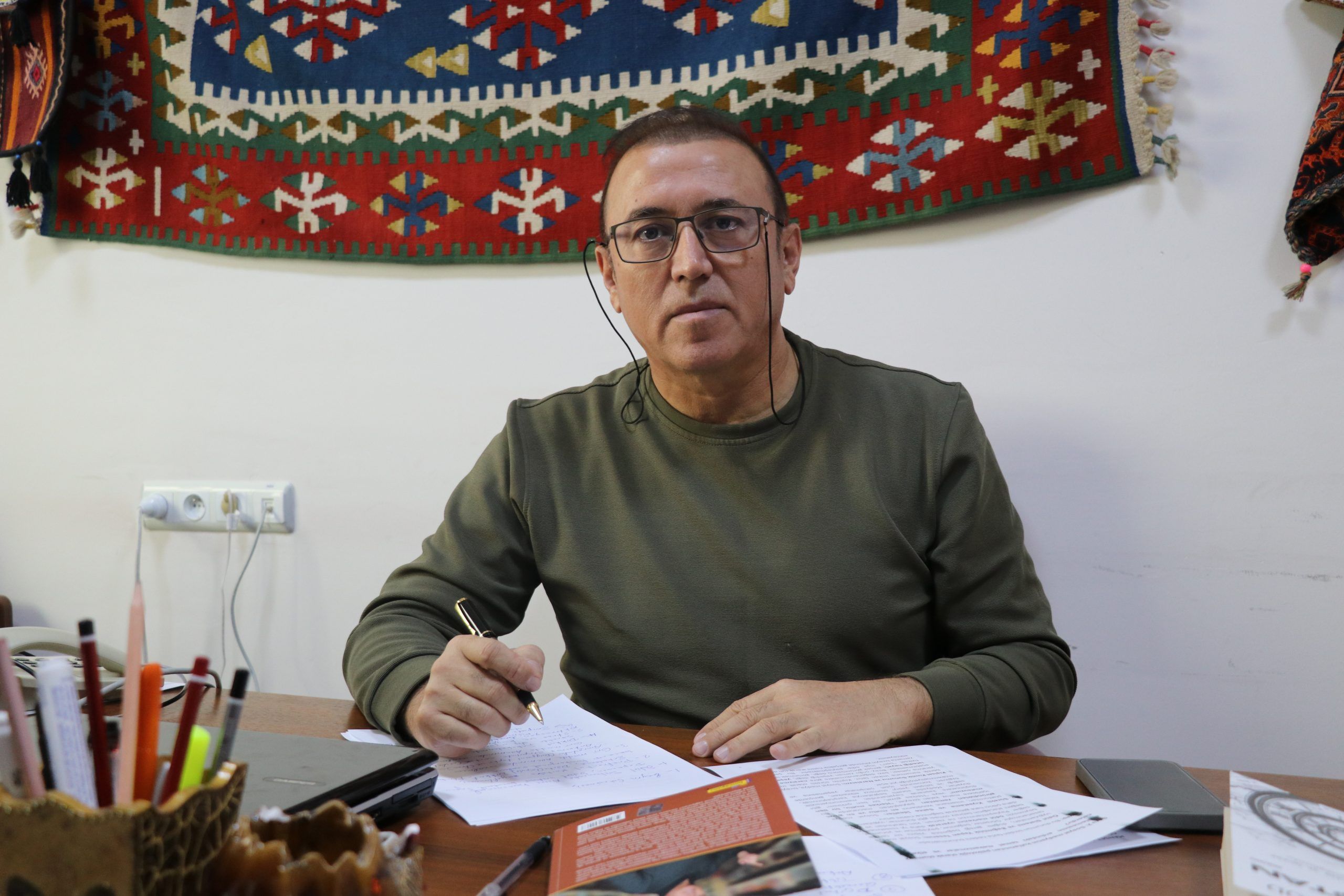 Gözlüklü bir kişi, masada kalemle yazı yazarken önünde kâğıtlar ve kitaplar bulunuyor.