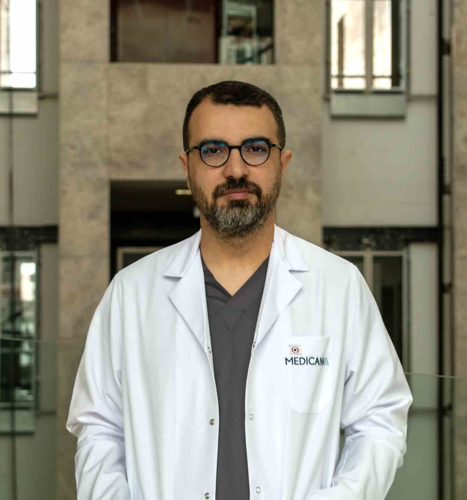 Gözlüklü ve sakallı bir kişi, üzerinde Medicana yazan beyaz önlükle bina girişinde kameraya bakıyor.