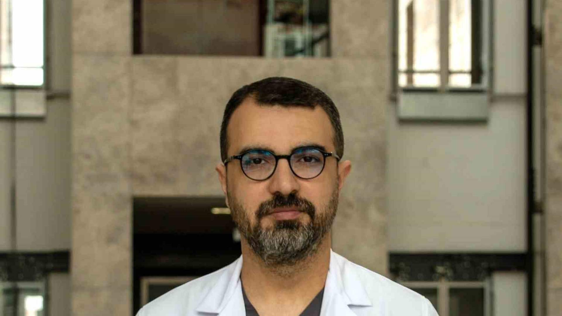 Gözlüklü ve sakallı bir kişi, üzerinde Medicana yazan beyaz önlükle bina girişinde kameraya bakıyor.