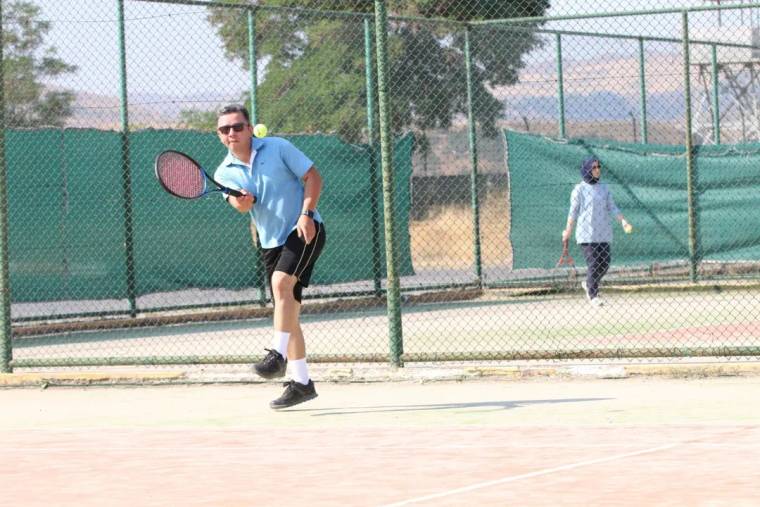 Açık tenis kortunda raket tutan bir erkek oyuncu topa vururken, arka planda başka bir oyuncu yürüyor.
