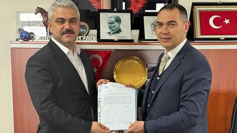İki erkek, Türk bayrakları ve portrelerin bulunduğu bir odada imzalı bir belgeyi birlikte tutuyor.