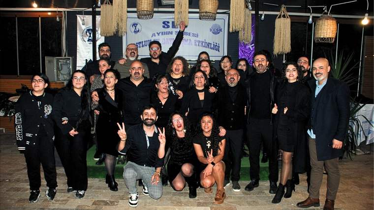 Kapalı bir mekânda, arkalarında dernek pankartı bulunan kalabalık bir grup birlikte poz veriyor.