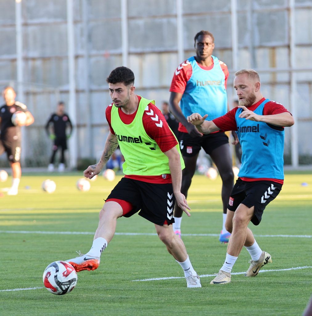 Antrenman sahasında yeşil yelekli ve mavi formalı iki futbolcu top mücadelesi sırasında koşuyor.