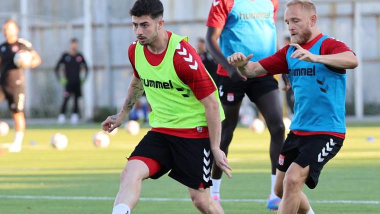 Antrenman sahasında yeşil yelekli ve mavi formalı iki futbolcu top mücadelesi sırasında koşuyor.