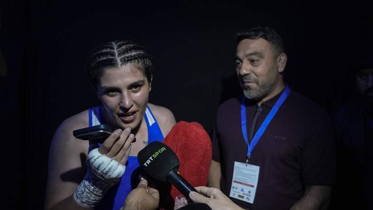Mavi formalı, elleri bandajlı bir sporcu, mikrofonlara konuşurken yanında yaka kartlı bir adam duruyor.