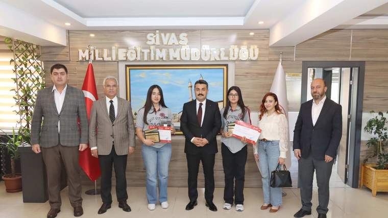Sivas Milli Eğitim Müdürlüğü yazılı salonda, ellerinde belge ve ödüller bulunan yedi kişi yan yana duruyor.