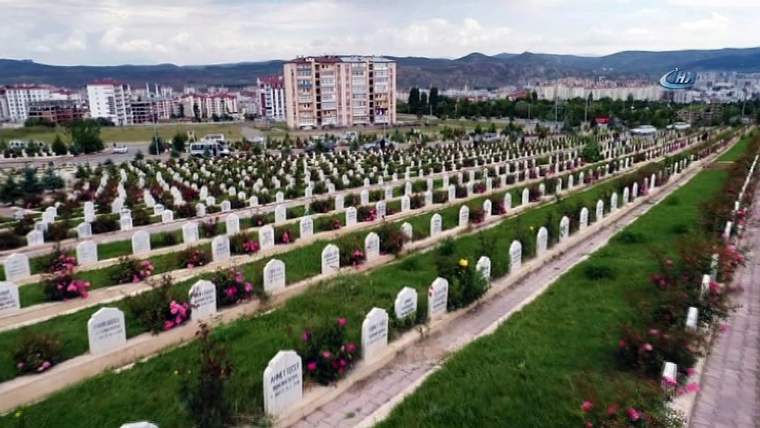 Yeşil alanlar ve çiçeklerle süslenmiş mezarların bulunduğu bir mezarlık görüntüsü.