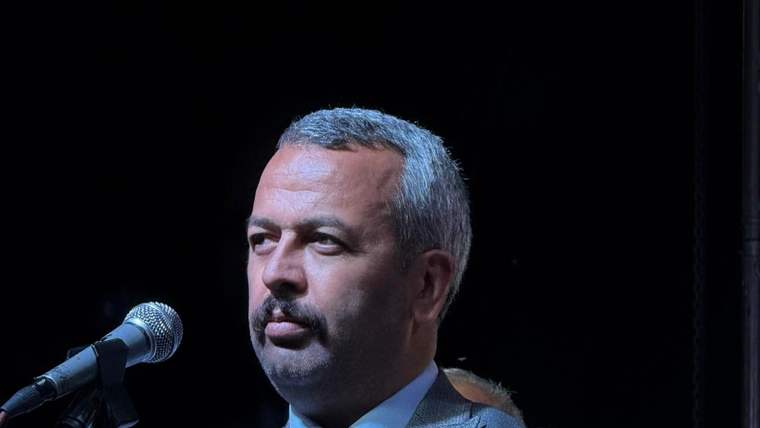 Kürsü mikrofonunun yanında takım elbiseli, kravatlı bir kişi sahnede otururken görülüyor.