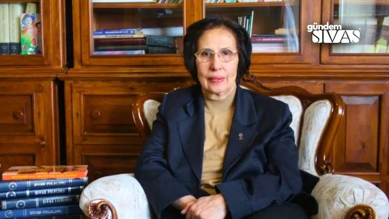 Bir kadın, kitapların bulunduğu bir rafın önünde, koltukta oturuyor.