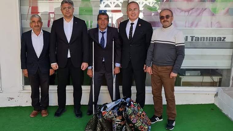 Bir iş yeri önünde beş erkek yan yana duruyor; öndeki renkli çantaların yanında bir kişi baston tutuyor.