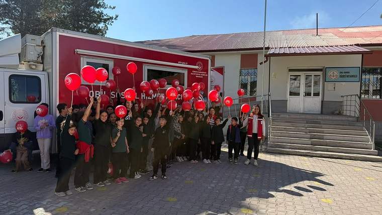 Kırmızı balonlar taşıyan çocuklar ve yetişkinler, kırmızı bir araç ile bina önünde birlikte duruyor.