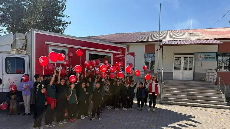 Kırmızı balonlar taşıyan çocuklar ve yetişkinler, kırmızı bir araç ile bina önünde birlikte duruyor.