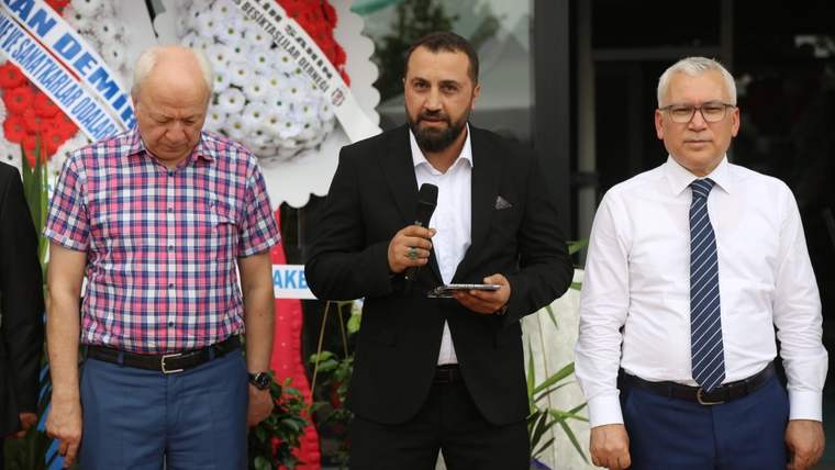 Kürsüde konuşan bir kişi, yanında iki kişiyle birlikte duruyor; arka planda çiçekler var.