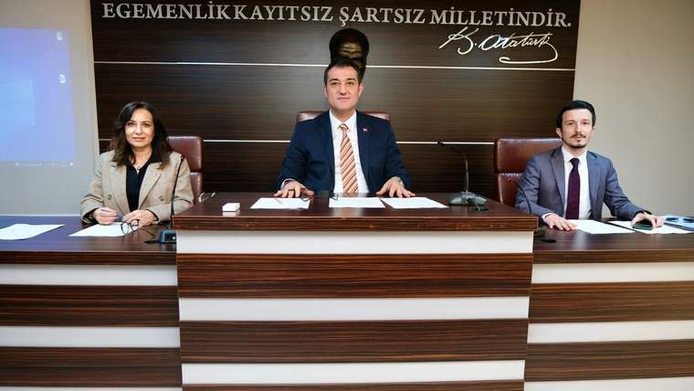 Toplantı salonunda üç kişi, üzerinde Atatürk imzası bulunan yazının önündeki kürsüde oturuyor.