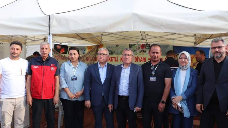 Fuar alanında, çeşitli kişiler bir arada poz veriyor; arka planda stantlar görünmekte.