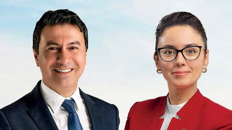 Kravatlı bir erkek ile kırmızı ceketli, gözlüklü bir kadın açık renkli arka plan önünde yan yana duruyor.