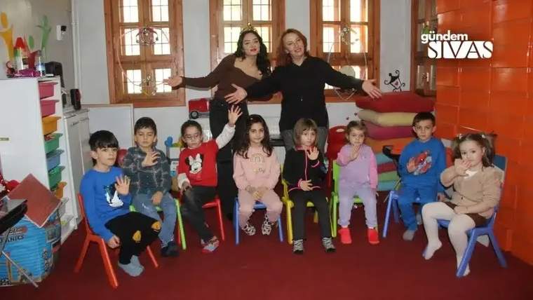 İki yetişkinin önünde oturan sekiz çocuk, ellerini havaya kaldırarak poz veriyor.
