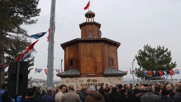 Ahşap yapılı bir saat kulesi, önünde kalabalık bir grup insan var.