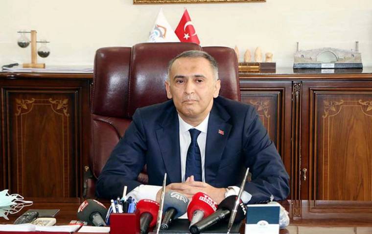 Masasında oturan bir kişi, önünde mikrofonlar ve Türk bayrağı var.