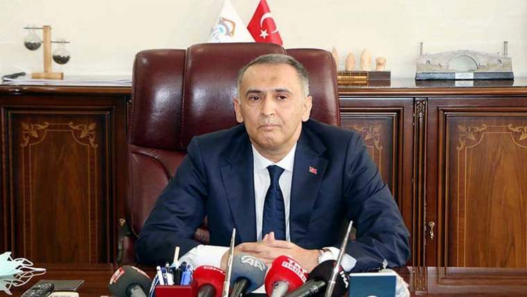 Masasında oturan bir kişi, önünde mikrofonlar ve Türk bayrağı var.