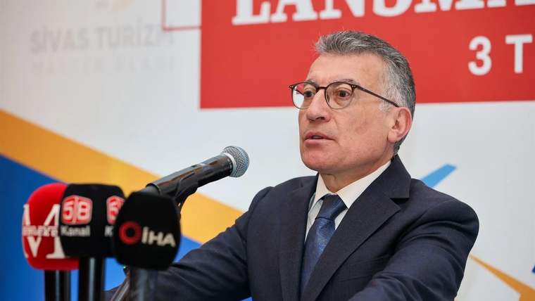 Kürsüde konuşan bir kişi, mikrofonun önünde duruyor ve arka planda tanıtım afişi var.