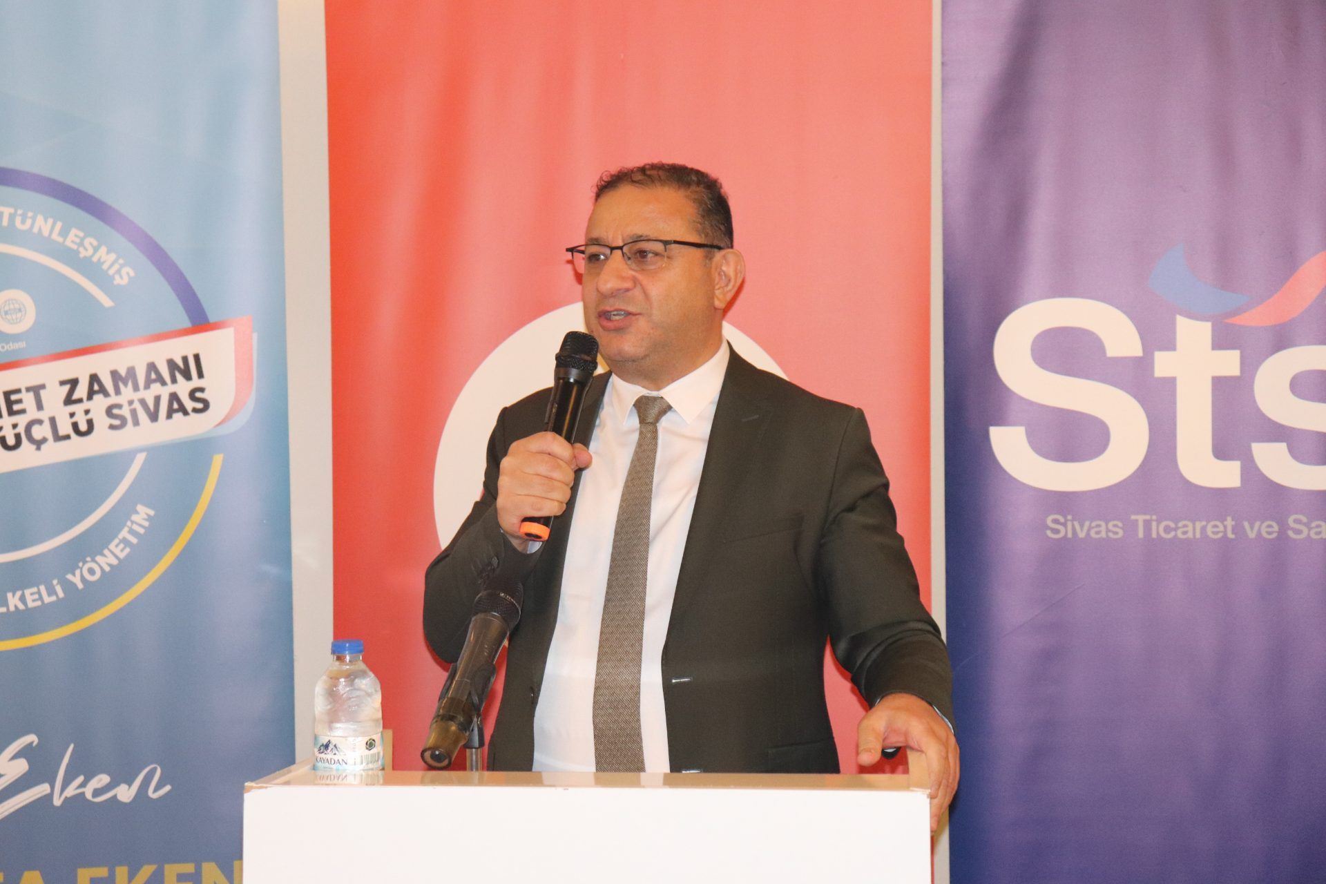 Kürsüde mikrofonla konuşan takım elbiseli bir kişi, arkasındaki pankartların önünde duruyor.