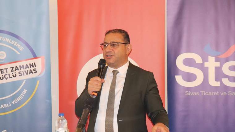 Kürsüde mikrofonla konuşan takım elbiseli bir kişi, arkasındaki pankartların önünde duruyor.