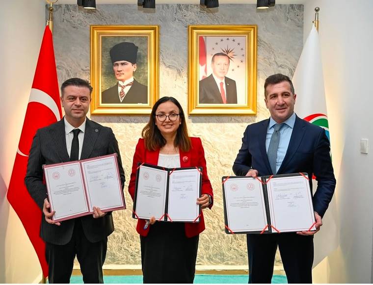 İç mekânda üç kişi, arkalarında Türk bayrağı ve portreler bulunan duvar önünde belgeler tutuyor.