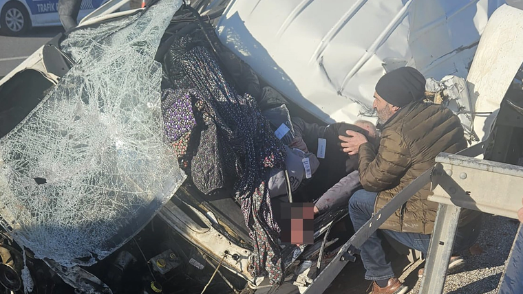 Devrilmiş bir aracın kırılmış ön camı ve ezilmiş kabini yanında, bere takan bir kişi içerideki yaralıya uzanıyor.