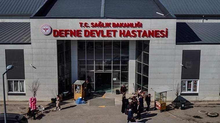 Üzerinde T.C. Sağlık Bakanlığı ve Defne Devlet Hastanesi yazan binanın girişinde insanlar görülüyor.