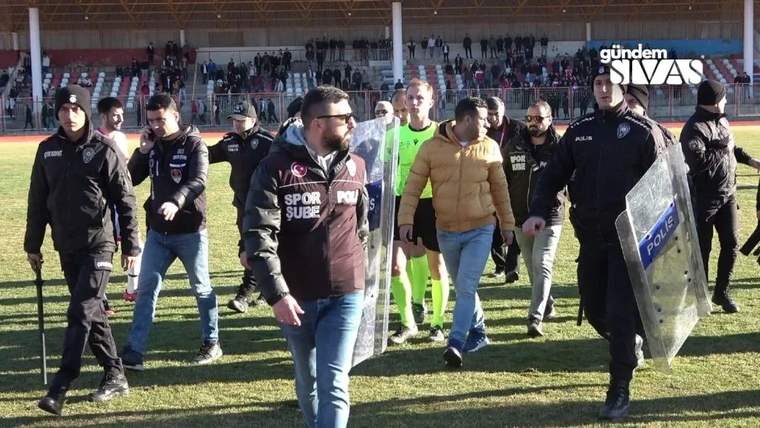 Futbol sahasında, spor yelekli bir adamın çevresinde polisler ve kalkan taşıyan bir görevli yürüyor.