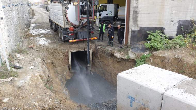 Dar bir kazı alanında kanalizasyon aracı su püskürtürken çevresinde üç görevli ve beton bir yapı görülüyor.