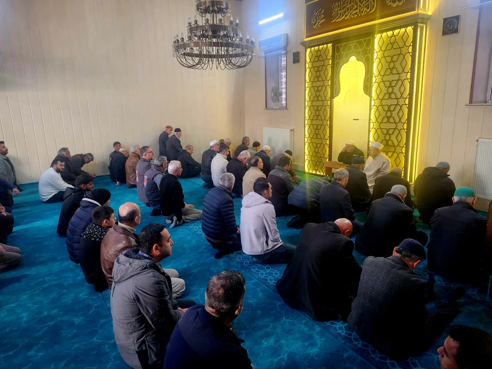 Cami içinde oturan kalabalık bir grup erkek, dua etmekte ve bir konuşmayı dinlemekte.