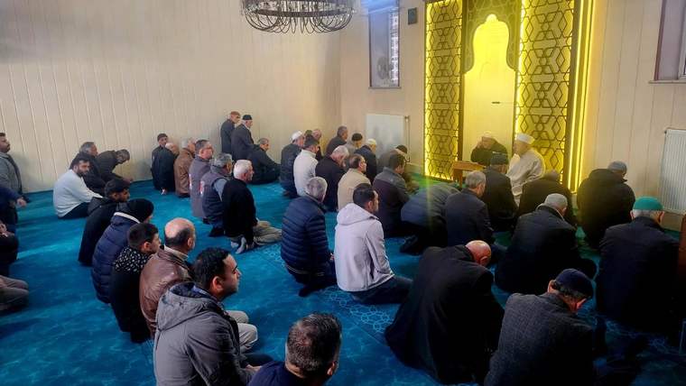 Cami içinde oturan kalabalık bir grup erkek, dua etmekte ve bir konuşmayı dinlemekte.