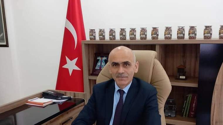 Takım elbiseli bir kişi, çalışma masasının arkasında oturuyor; yanında Türk bayrağı ve raflar bulunuyor.