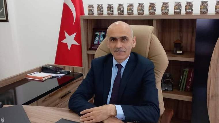 Kravatlı bir kişi, Türk bayrağı ve raflarda çeşitli nesnelerin bulunduğu ofiste masada oturuyor.