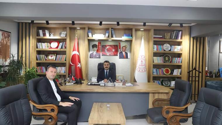 Bir ofiste masanın arkasında oturan iki erkek, Türk bayrakları ve kitaplıkların önünde görülüyor.