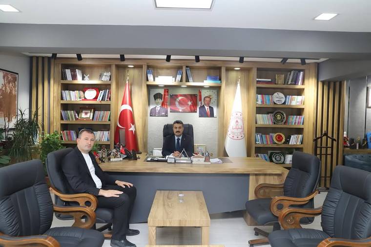 Bir ofiste masanın arkasında oturan iki erkek, Türk bayrakları ve kitaplıkların önünde görülüyor.