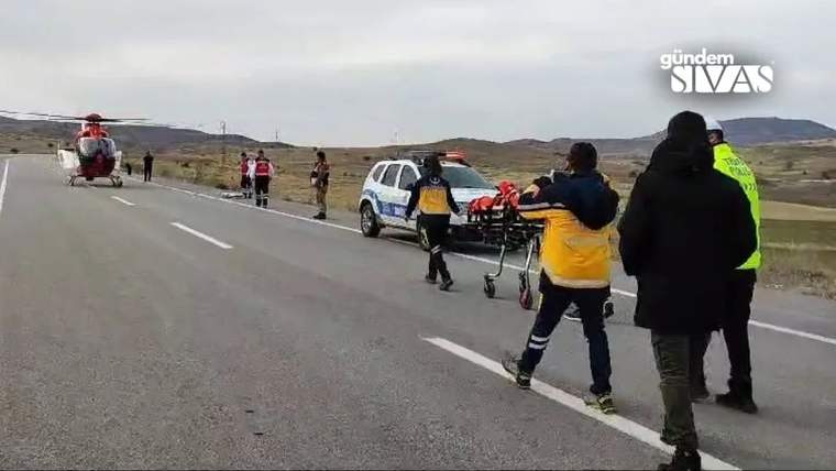 Karayolunda sağlık görevlileri sedye taşırken, ileride bir helikopter ve iki araç görülüyor.