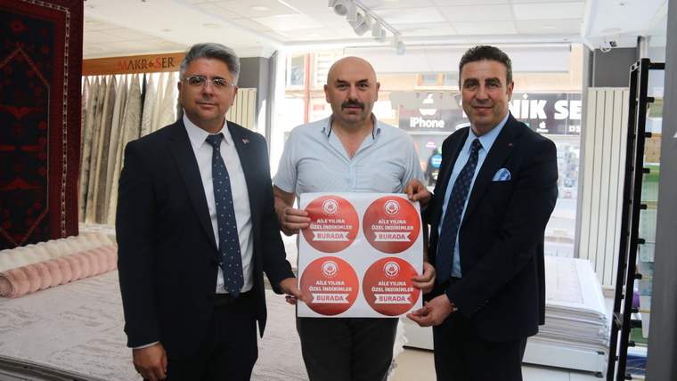 Üç kişi, elinde indirim afişi tutarak gülümsüyor. Arka planda yataklar ve ev tekstili ürünleri var.