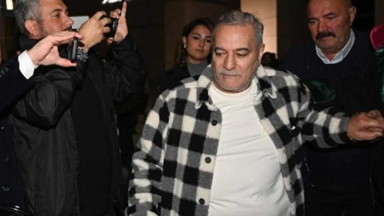 Kareli bir kaban giymiş bir adam, kalabalık bir grup arasında yürürken görünüyor.