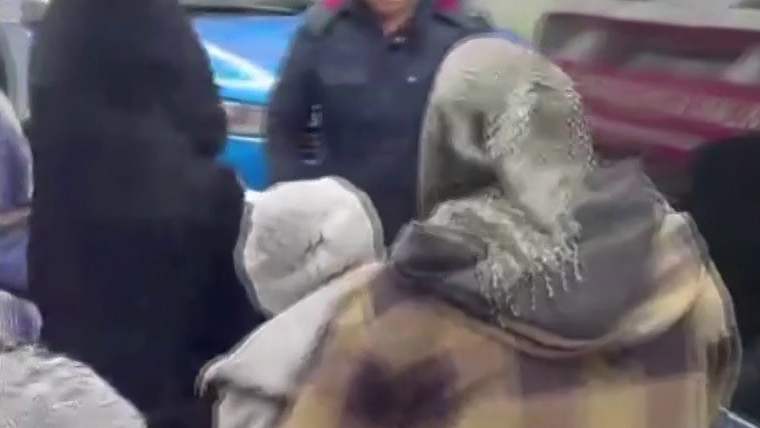 Başörtülü ve battaniyeye sarınmış bir kişi, çevresindeki iki kişiyle birlikte kapalı alanda görülüyor.