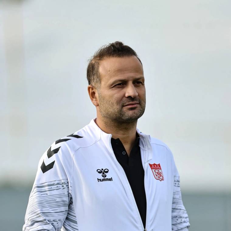 Beyaz spor üstü giyen bir adam, üzerinde Sivasspor arması bulunan kıyafetle dışarıda duruyor.