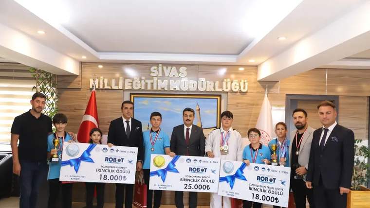 Sivas Milli Eğitim Müdürlüğü yazılı salonda bir grup öğrenci ve yetişkin, madalya ve ödül çekleriyle duruyor.