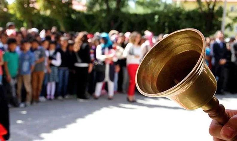 Bir grup öğrenci ve yetişkin, okul bahçesinde toplanmış; ön planda bir çan yer alıyor.